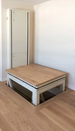 A Telecab through the floor lift with a light wood surface and a enclosed structure against a white support wall.