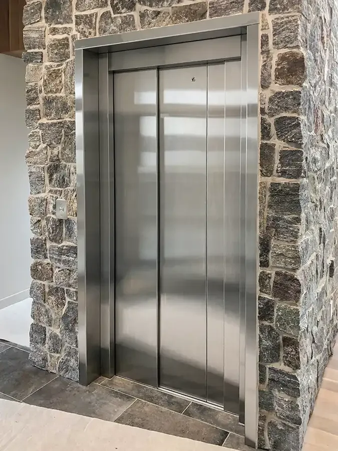 Residential elevator with stainless steel sliding doors framed by natural stone, installed in a modern upscale home interior.