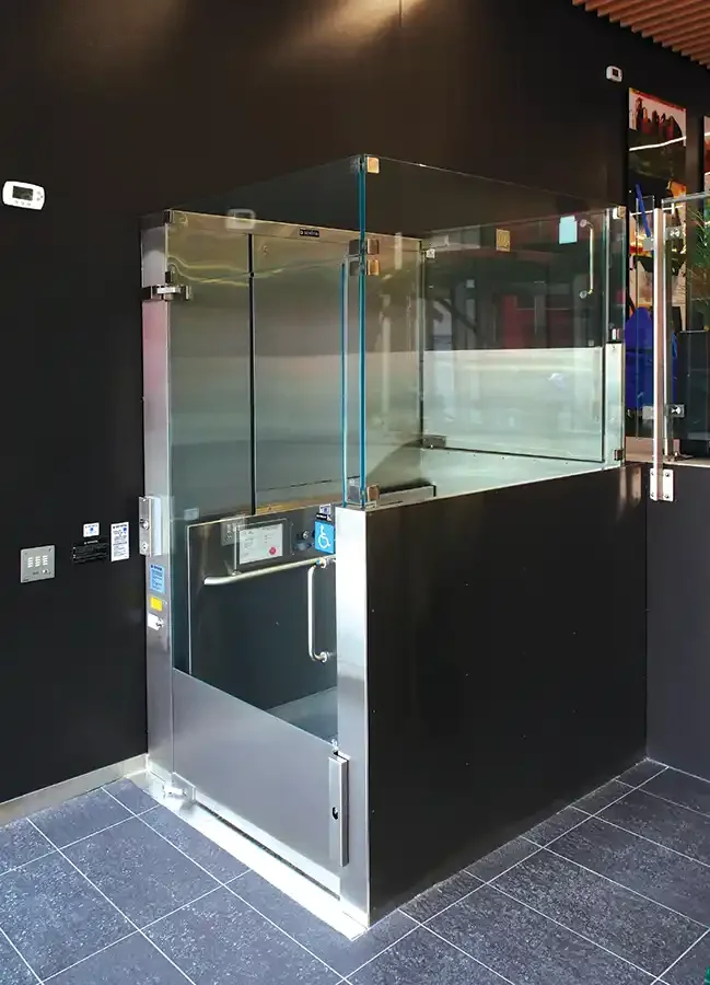 Glass-enclosed commercial platform lift with stainless steel base and transparent panels set against a dark interior wall