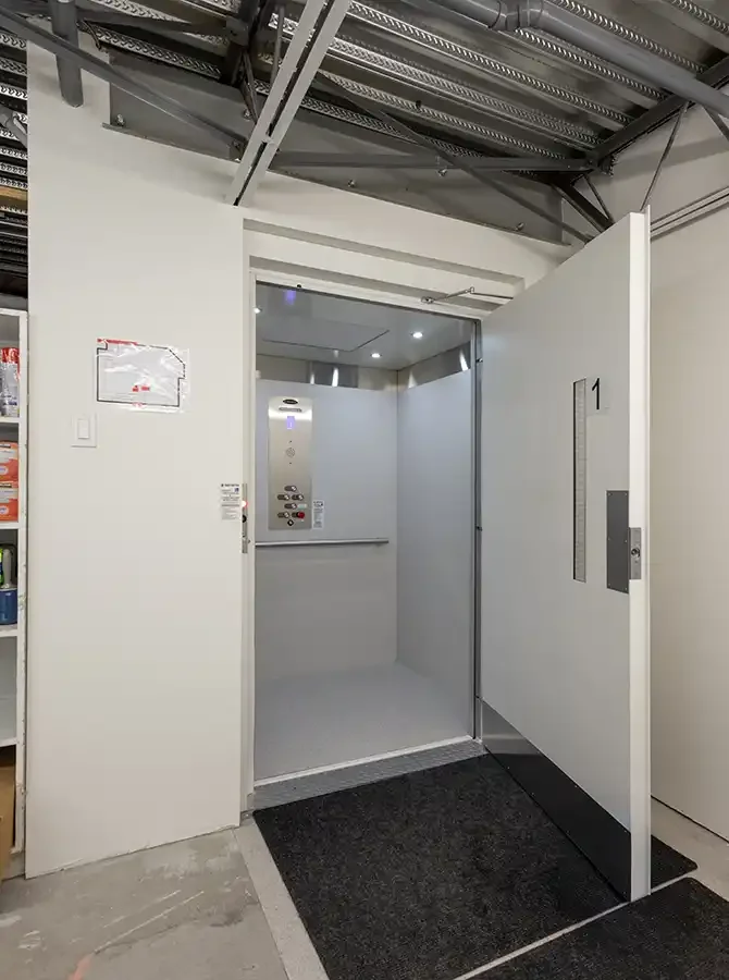 Open vertical platform lift behind a commercial service door, with clean white interior panels and control panel visible