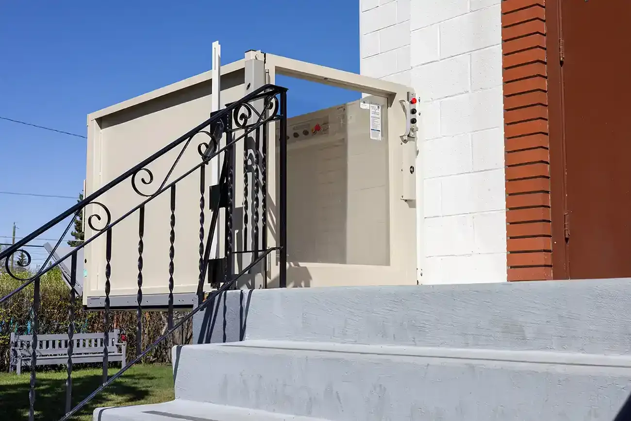 Outdoor vertical platform lift with beige metal enclosure installed beside a residential building, providing wheelchair and mobility accessibility from ground level to elevated entry with exterior stairs and railing.