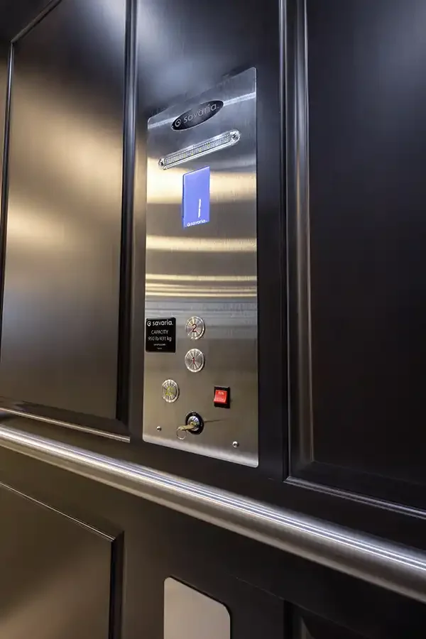 Residential elevator interior featuring black‑colored hardwood wall panels, stainless steel control panel, illuminated floor display, handrail, and luxury modern design.