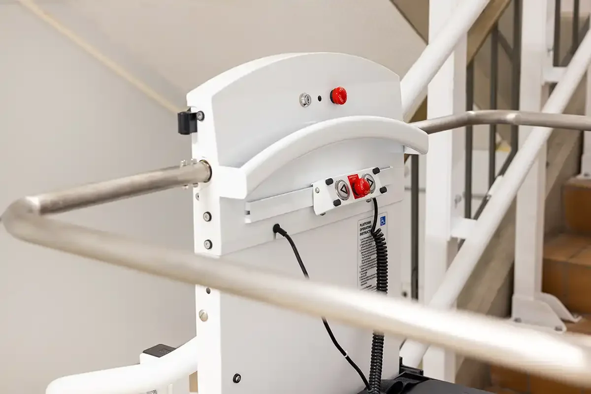 Close-up of inclined platform lift control head with safety arms, emergency stop button, and handrails mounted beside a commercial staircase