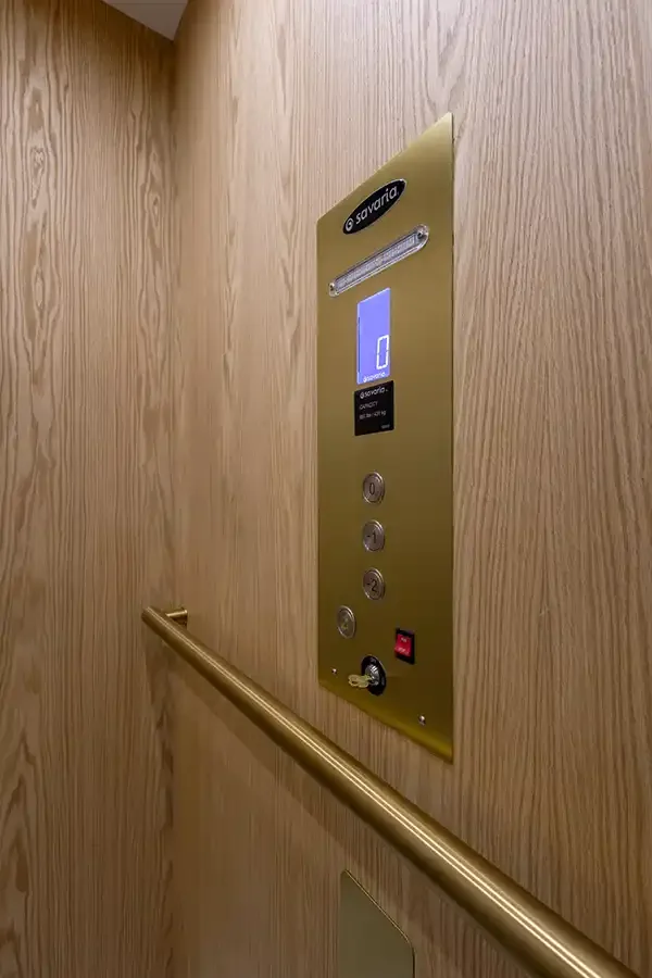 Residential elevator control panel with brass‑finish faceplate, Savaria branding, digital floor display, illuminated buttons, emergency stop, key switch, and brass handrail on wood‑paneled cabin wall.