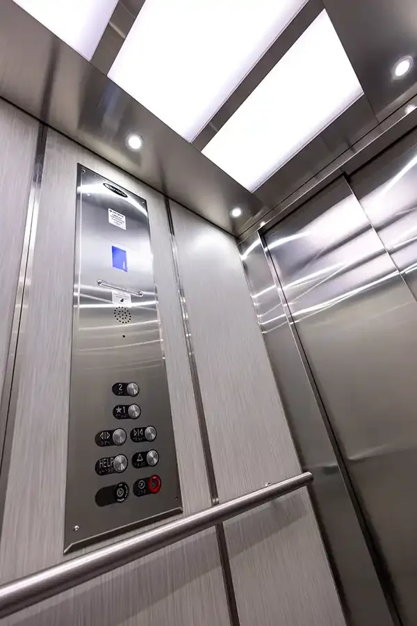 Residential elevator interior with stainless steel control panel, illuminated ceiling panels, and brushed metal walls