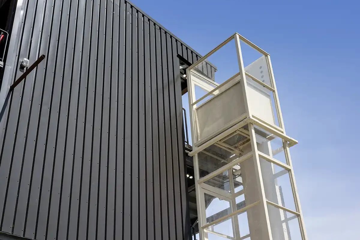 Outdoor vertical platform lift tower attached to a modern building exterior with glass panels and metal framing