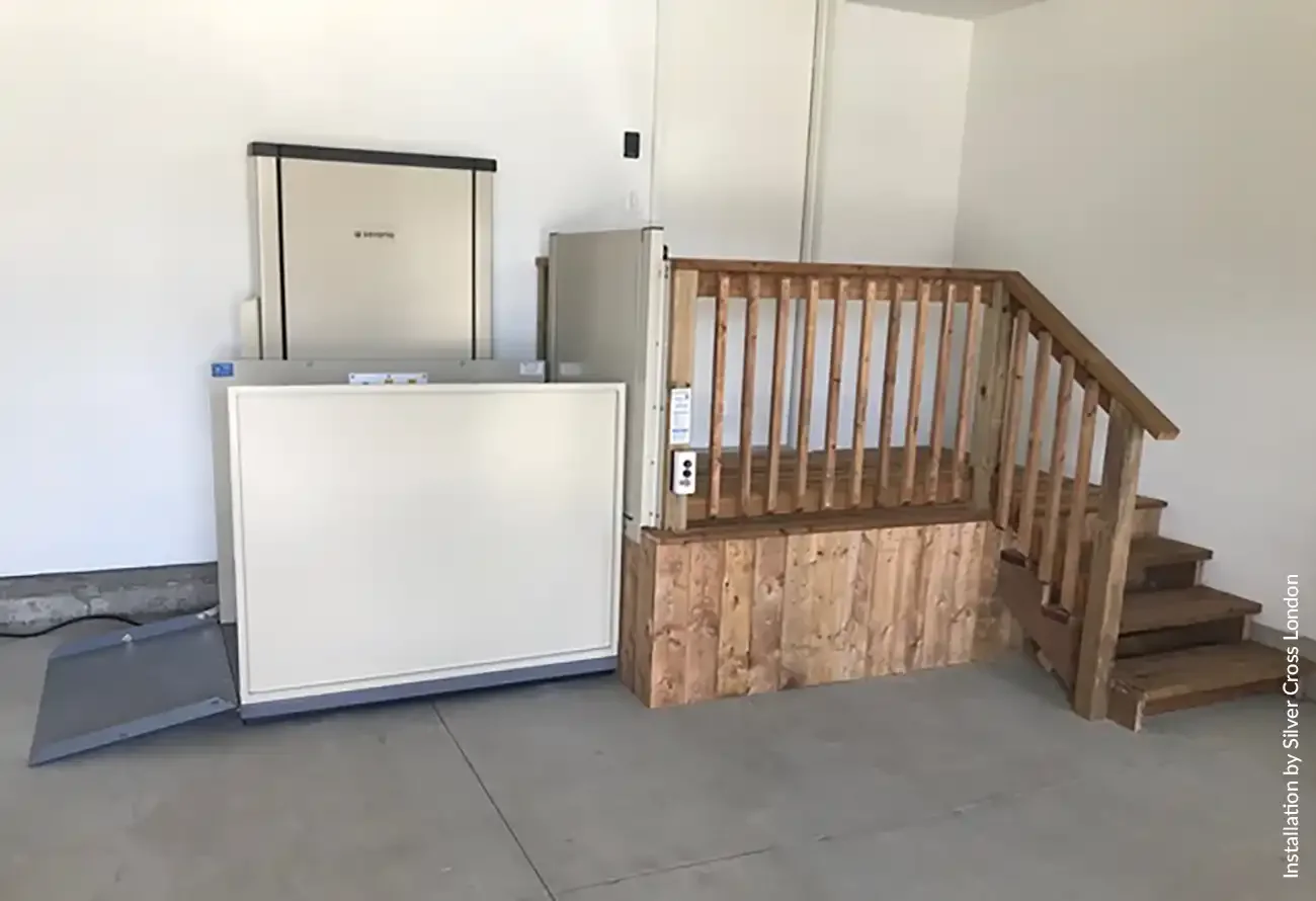 Indoor residential vertical platform lift with beige metal enclosure installed in a garage or interior space, providing wheelchair and mobility accessibility to an elevated landing with adjacent wooden stairs and railing.