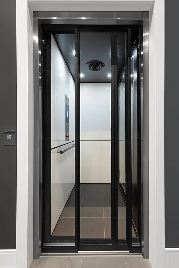 Glass-front residential elevator with black framing, white interior panels, and recessed ceiling lights