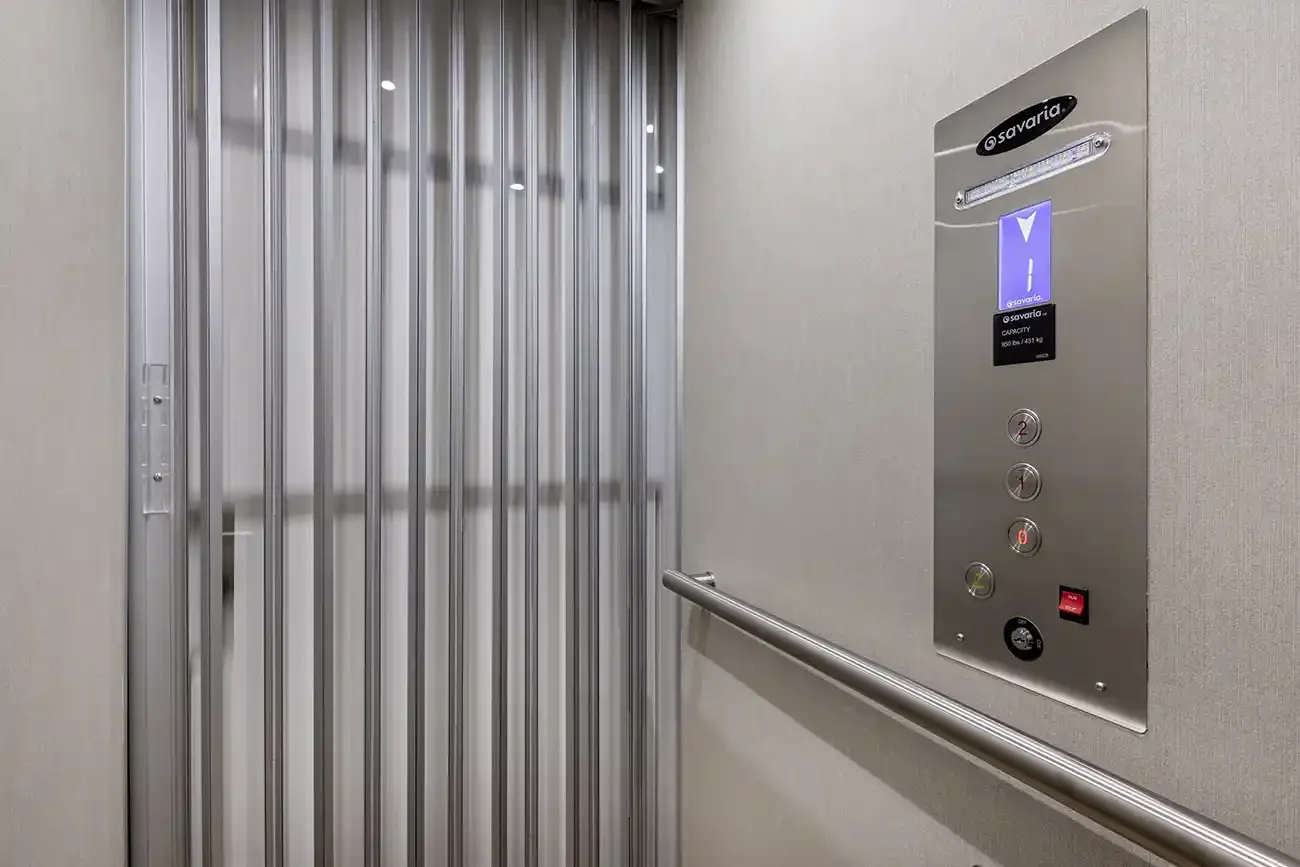 Residential elevator interior featuring a silver accordion safety gate, stainless steel control panel with digital floor display, horizontal handrail, and modern neutral wall finishes.