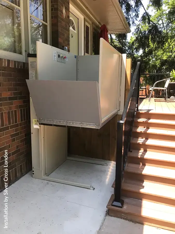 Outdoor residential vertical platform lift with beige metal enclosure installed beside a brick home, providing wheelchair and mobility accessibility between ground level and an elevated wooden deck next to exterior stairs.