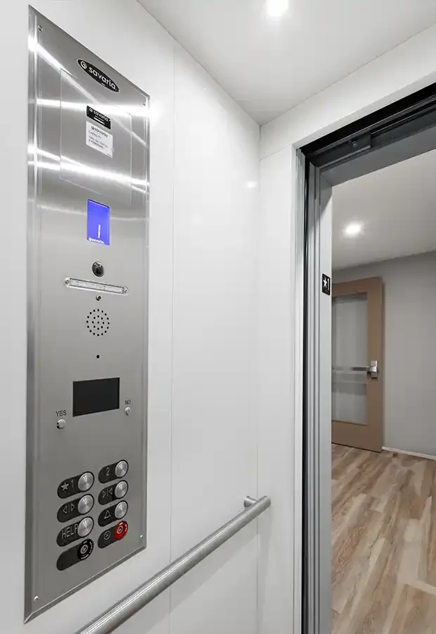 Commercial elevator interior with stainless steel control panel, handrail, and open door leading to a modern hallway