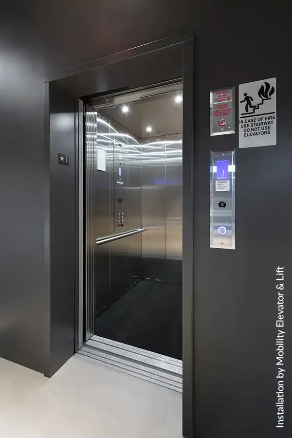 Open commercial elevator with stainless steel interior, illuminated ceiling panels, and dark corridor walls