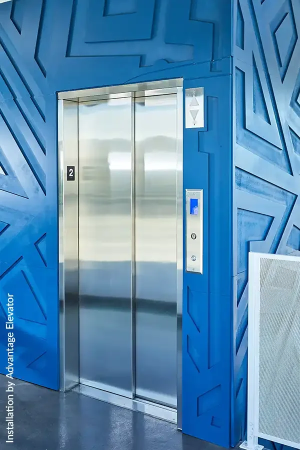 Closed stainless commercial steel elevator doors set into a bold blue geometric wall with a digital call panel