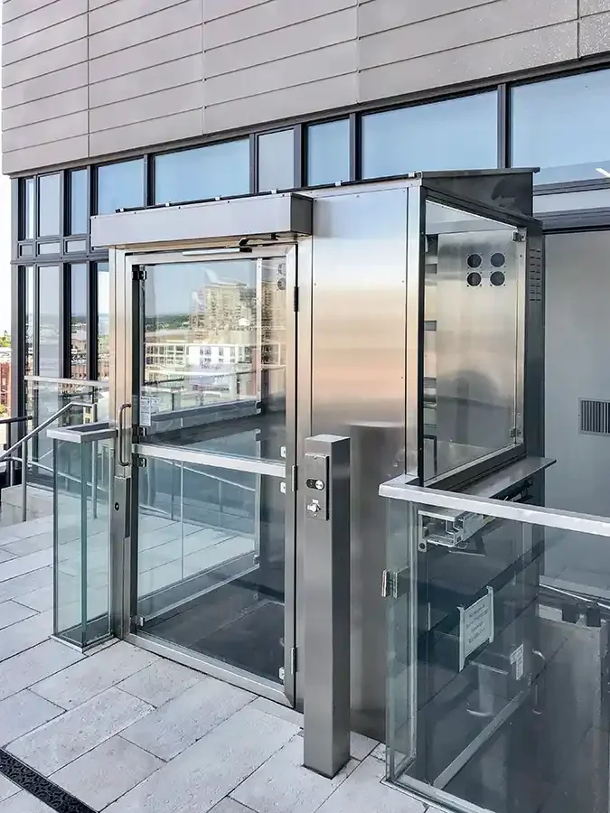 Glass-enclosed wheelchair platform lift installed on an outdoor terrace beside a modern building façade