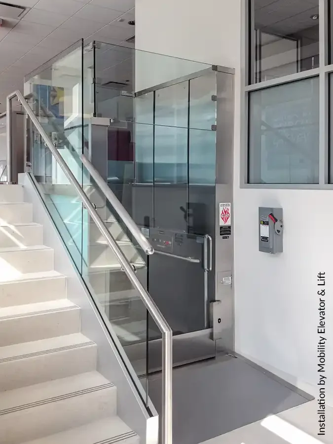 Glass-enclosed commercial platform lift beside a stairway in a bright, modern interior with stainless steel railings