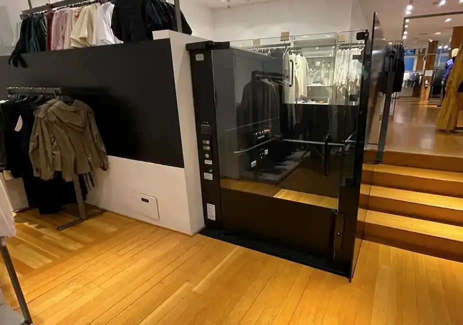 Glass-enclosed commercial platform lift integrated into a retail store beside a short set of wooden steps