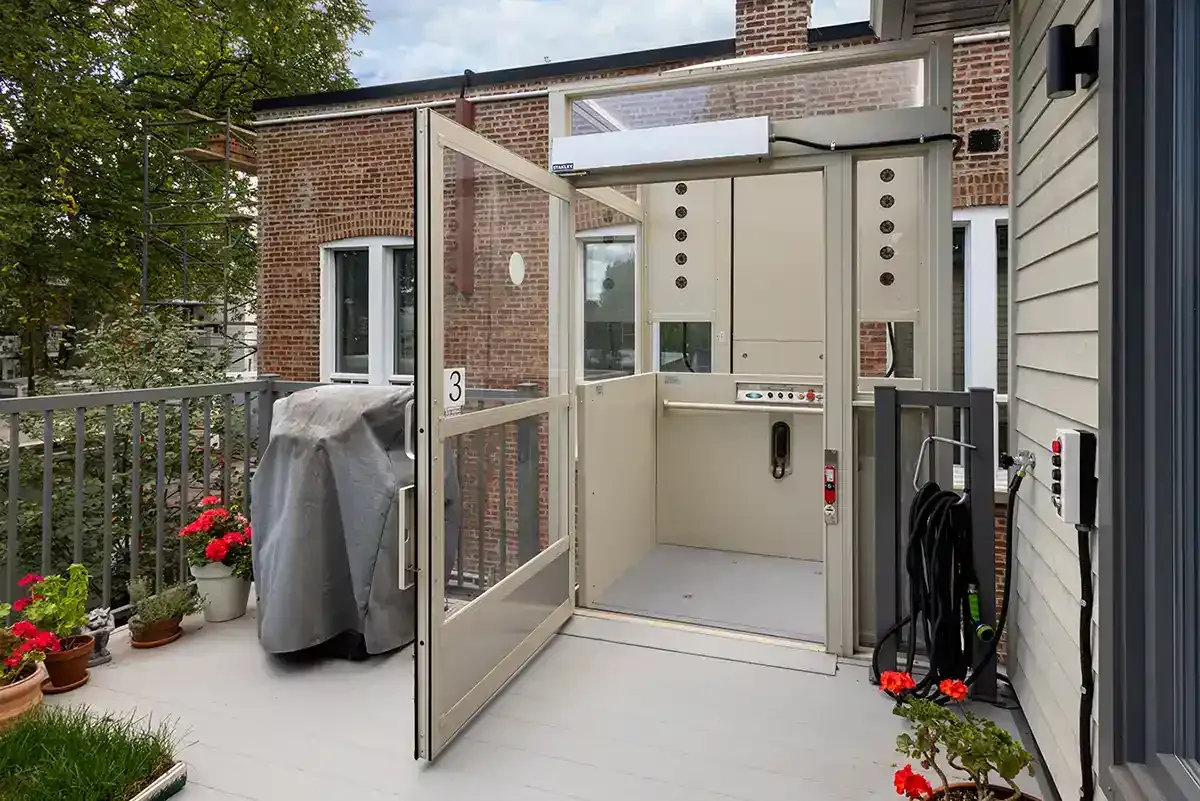 Outdoor wheelchair vertical platform lift with open door on a residential balcony beside a brick building