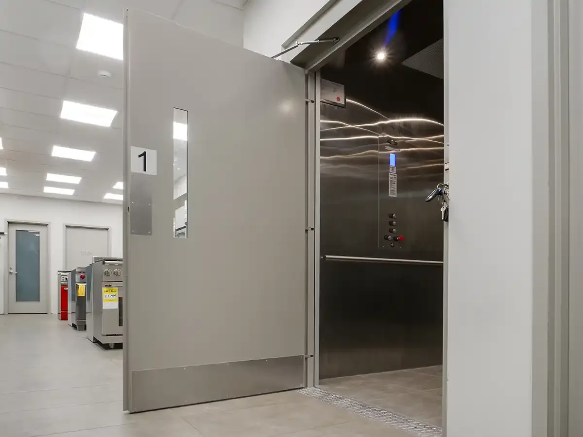 Open vertical platform lift behind a commercial door, featuring stainless steel interior panels, control panel, and recessed ceiling lights