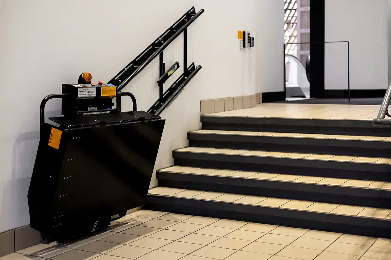 Folded inclined platform lift parked beside a commercial staircase, showing compact wall‑mounted rail system and minimal obstruction when not in use.
