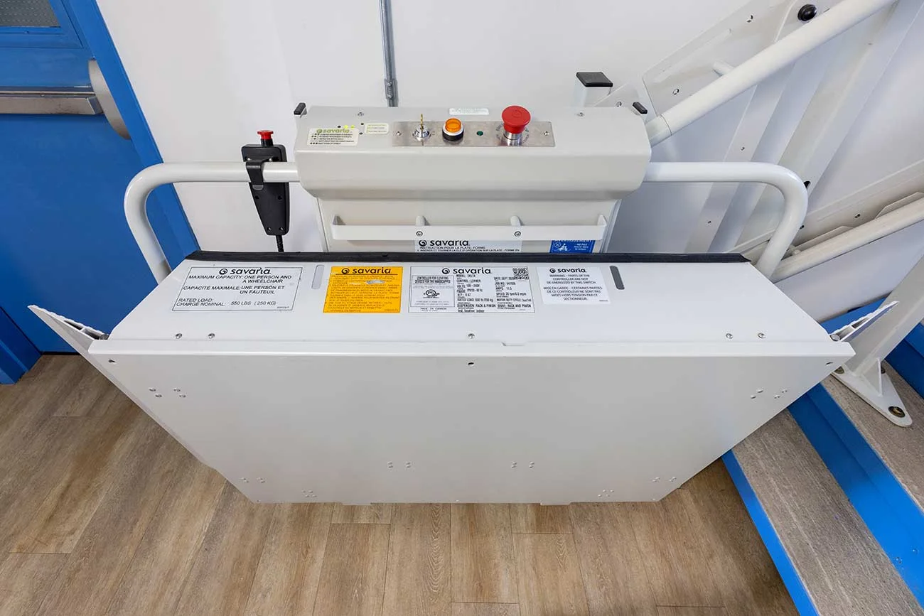 Savaria Delta inclined platform lift drive unit and control assembly with emergency stop buttons, safety arms, and fold‑up wheelchair platform installed on an indoor staircase.