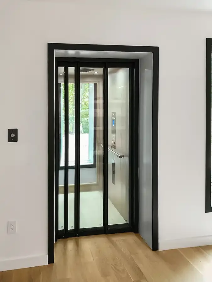Residential elevator with black‑framed glass sliding doors, stainless steel interior wall, control panel, and wood flooring, installed in a modern home hallway.