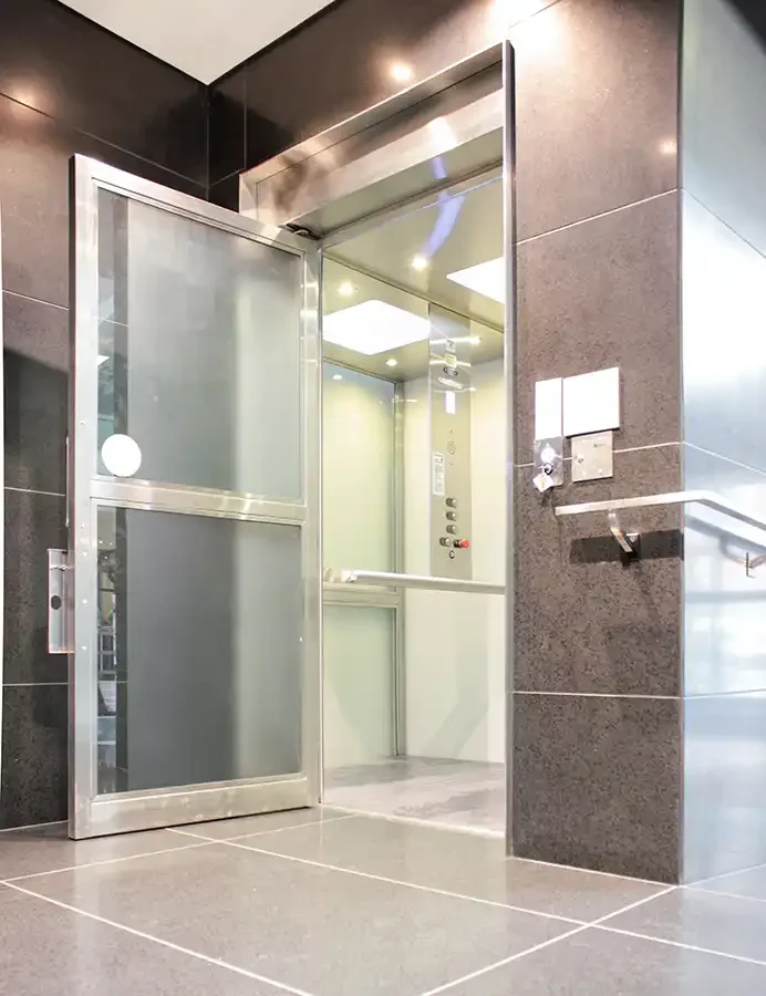 Glass-enclosed vertical platform lift with transparent door, stainless trim, and illuminated interior in a commercial lobby