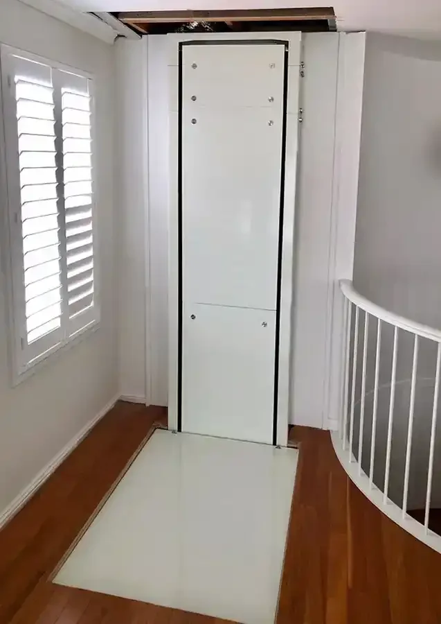 Through‑floor residential lift with white flush panels and landing platform installed in an upper hallway with wood flooring