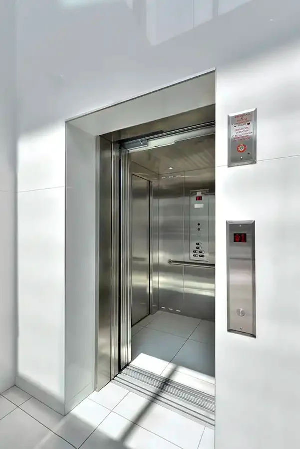 Open stainless steel elevator with bright tiled interior and digital call panel on a white wall