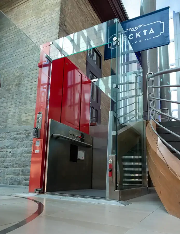 Glass-enclosed commercial elevator with red interior structure beside a curved staircase in a modern building atrium