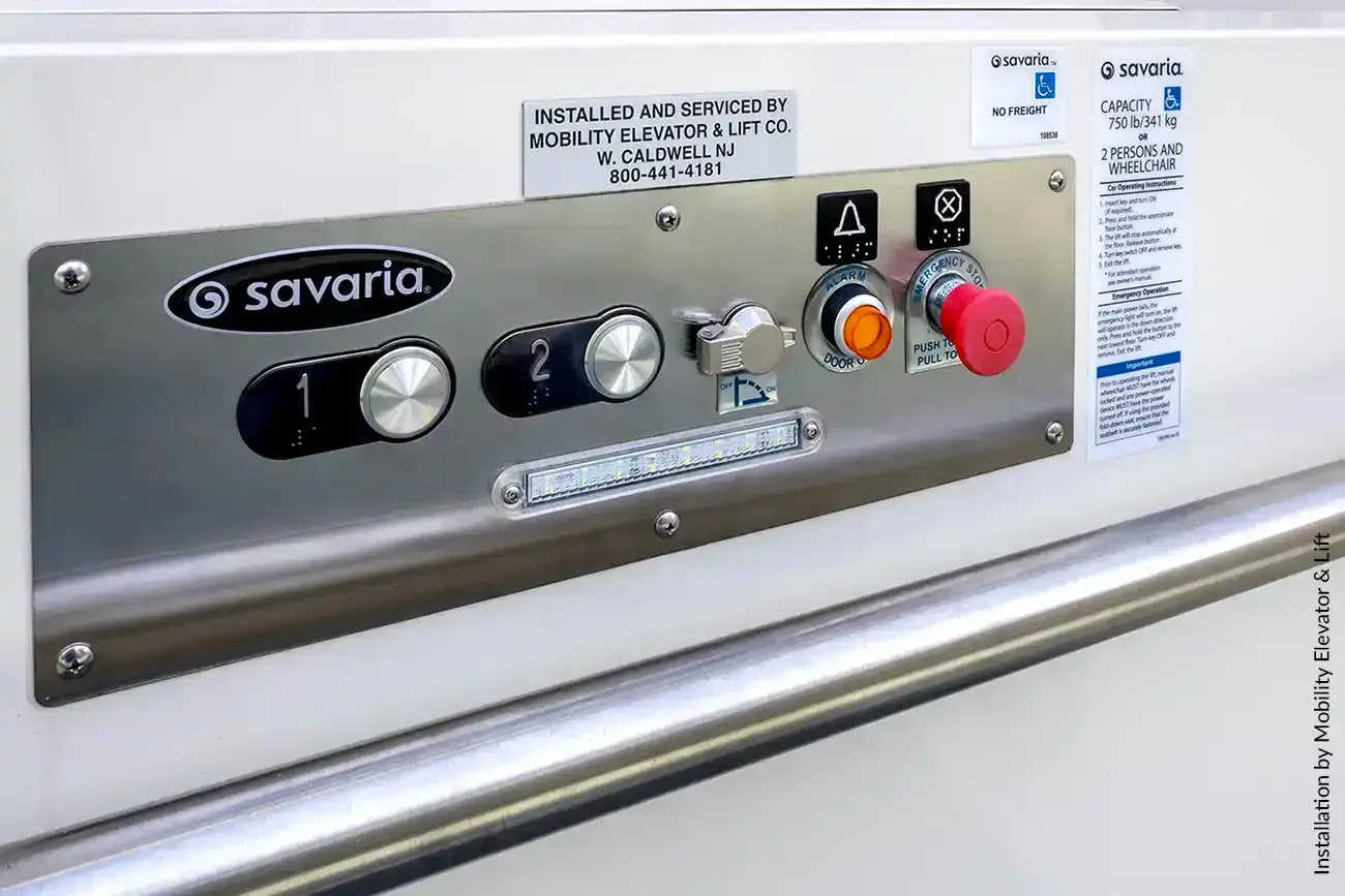 Close-up of a Savaria platform lift control panel with floor buttons, emergency stop, alarm, and operating instructions