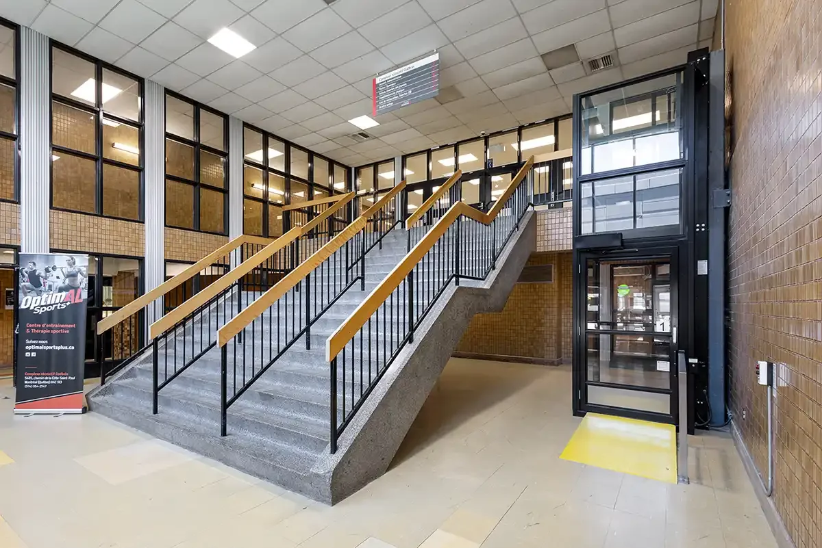 Wide commercial staircase with handrails beside a glass-enclosed vertical platform lift in a public building lobby