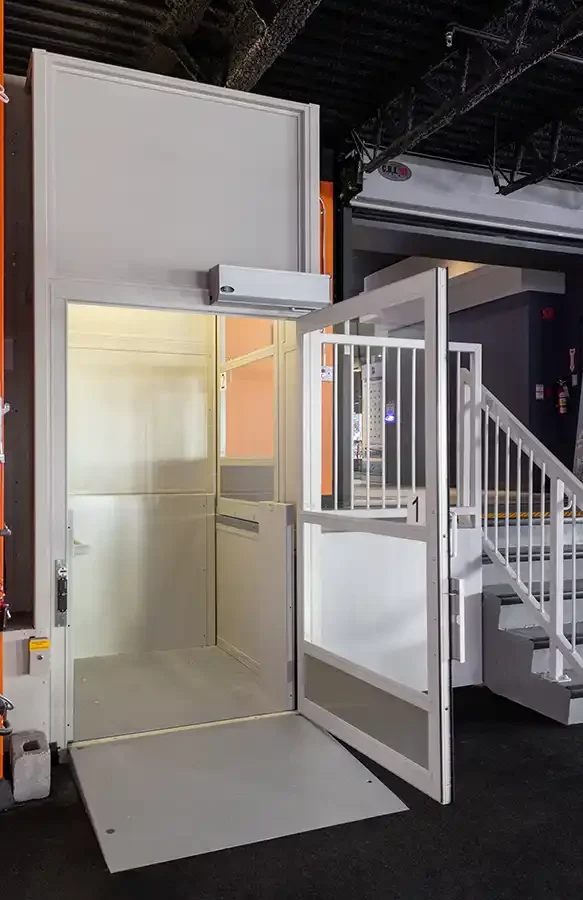 Open vertical platform lift with white interior and swing door beside an industrial staircase in a commercial setting
