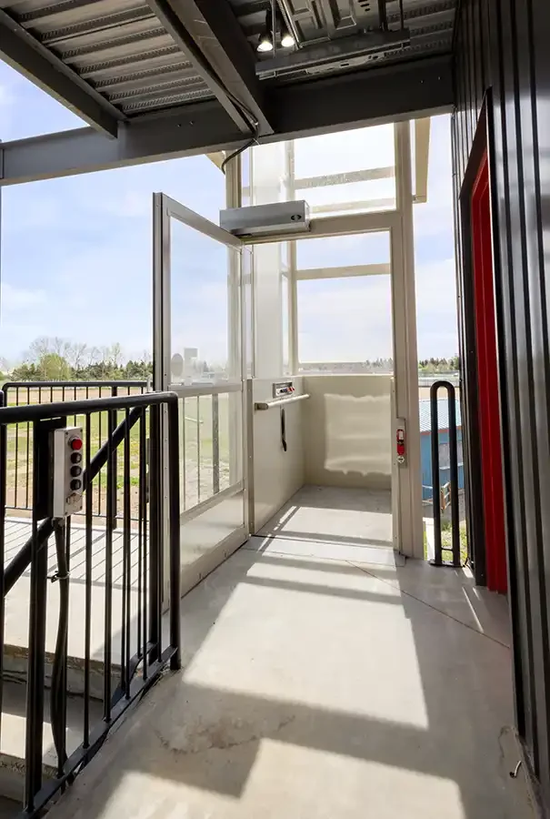Open vertical platform lift with white interior and glass door at an outdoor commercial landing under a covered structure