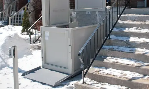 A unenclosed vertical platform lift installed next to a staircase covered in snow and ice.