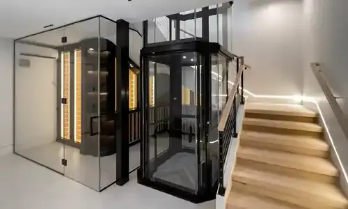 An octagonal panoramic winding drum elevator stationed at the bottom landing, next to a brightly lit staircase.