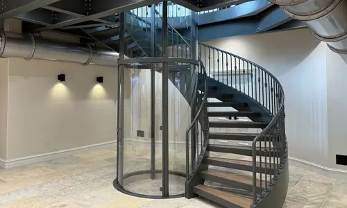 A round glass winding drum elevator installed in the center of a spiral curved staircase.