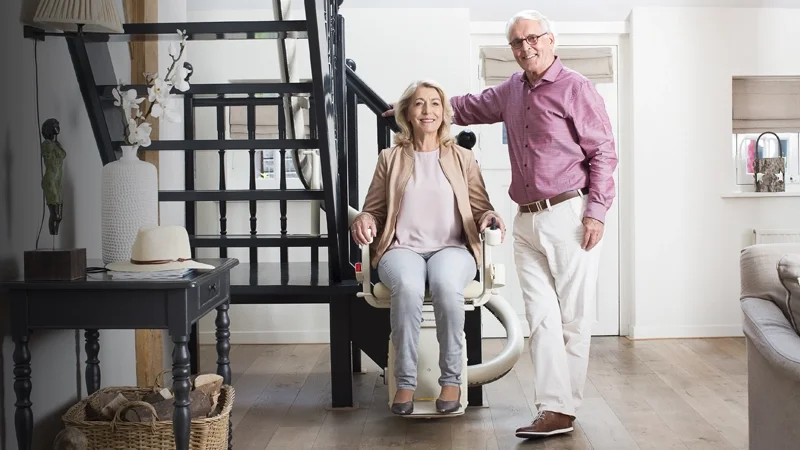 A smiling couple riding a single-rail curved stairlift along a black open staircase inside their home.