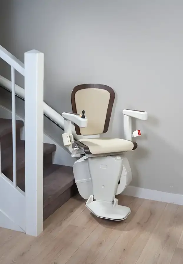 Stairlift with padded seat, armrests, and fold-down footrest positioned at the base of a straight residential staircase with carpeted steps and a white railing.
