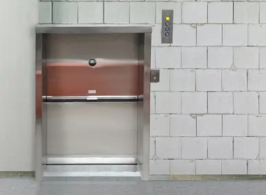 Material lift with a stainless‑steel vertical sliding door installed in a block‑wall setting, featuring a control panel with illuminated call buttons mounted to the right of the opening.