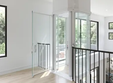 Panoramic glass residential elevator at an upper floor landing, featuring a cylindrical glass cabin with open safety gate, black railing, and bright modern interior with natural light.