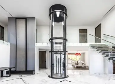Glass cylindrical residential elevator positioned in the center of a bright modern foyer, featuring transparent curved panels, a black frame, and a marble‑pattern floor beneath it, with a floating glass‑railed staircase to the right and open living and dining areas in the background.