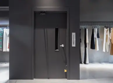 Vertical reciprocating conveyor shown in a retail back‑of‑house setting, with a dark swing door featuring a narrow vision panel, a wall‑mounted control panel to the right, and clothing racks visible on both sides of the space.