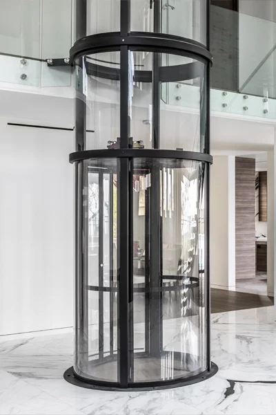 Round panoramic glass elevator with a black frame installed in a modern home interior with marble flooring and glass railings.