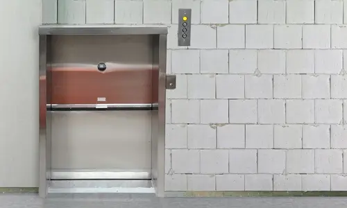 Stainless‑steel dumbwaiter elevator with a small service door set into a concrete block wall, shown with a wall‑mounted control panel.