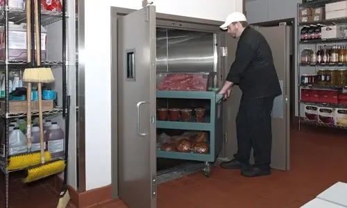 A worker in a commercial kitchen loads a cart of food items into a stainless‑steel material lift.