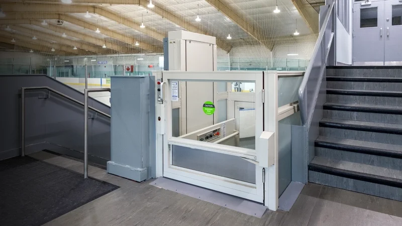 White-framed, unenclosed vertical platform lift installed next to a flight of stairs in a hockey arena.