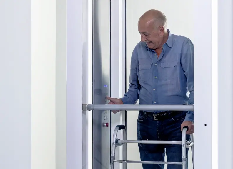 Person standing inside a Luma through‑the‑floor lift, using a walker and pressing the control panel inside the enclosure.