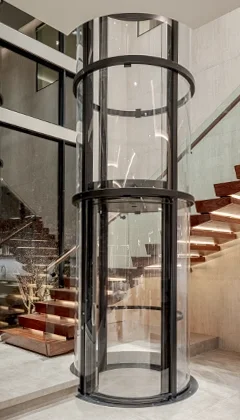 Glass cylindrical home elevator with black framing installed beside a modern open staircase.