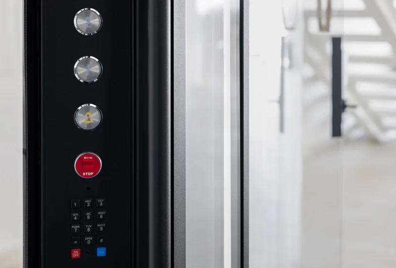 Elevator cab operating panel with three round metal buttons above a red stop button, followed by a numeric keypad and function keys, all mounted beside a glass elevator door.