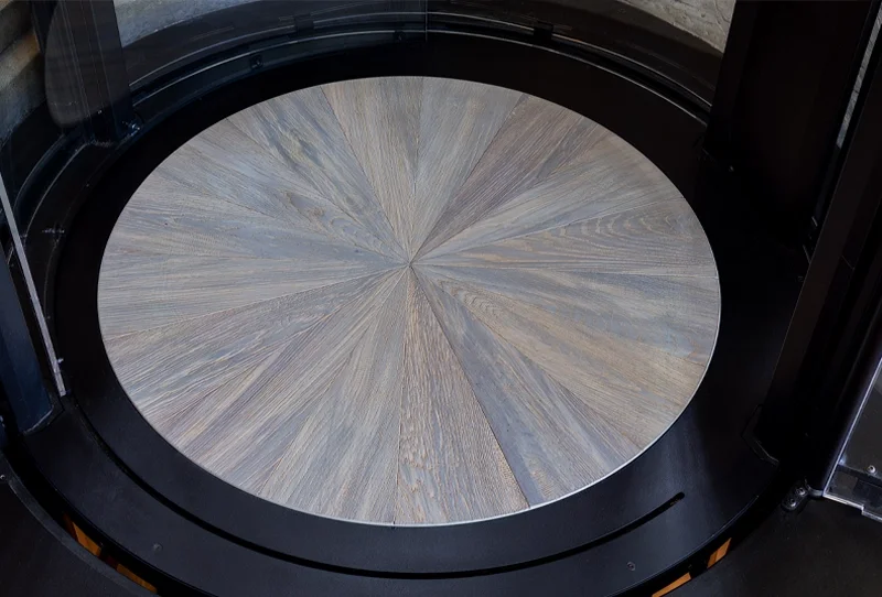 Circular elevator cab floor featuring a radial wood‑pattern design set within a black circular frame at the base of a glass‑walled home elevator.